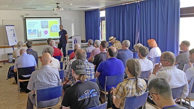 Ein Vortrag in einem Innenraum mit vielen Sitzplätzen. Im Vordergrund steht ein Sprecher vor einer Präsentation, während das Publikum aufmerksam zuhört. Die Gruppe setzt sich aus Männern und Frauen unterschiedlichen Alters zusammen.