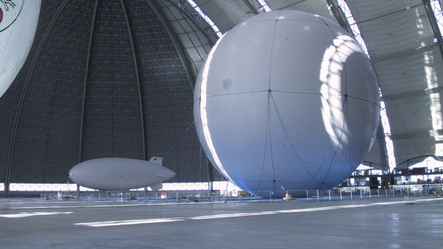 Ein großer, weißer Ballon einem kleinen, weißen  Luftschiff. Alles in einer großen Halle mit gewölbter Decke. Das Licht fällt durch die Fenster und erzeugt Schatten auf dem glatten Boden.