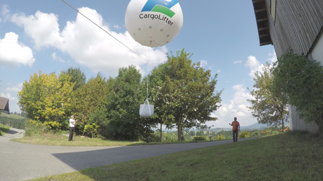 Eine großer Balllon dem Logo von CargoLifter schwebt über einer Wiese, rechts und links ein Führungsseil. Es hängt ein weißer Behälter darunter. Im Hintergrund sind grüne Bäume und ein Gebäude zu sehen, während zwei Personen, eine stehend und eine laufend, im Vordergrund stehen. Der Himmel ist klar mit einigen Wolken.